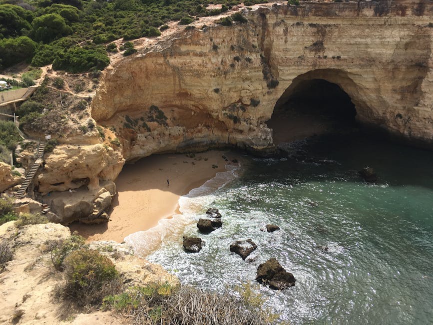 A scenic coastal landscape featuring a small, sandy beach nestled within a natural limestone cliff formation. The cliff has a prominent arch-shaped opening, with a textured surface displaying hues of cream, ochre, and light brown, along with darker patches of weathered rock. The sandy beach area is partially shaded by the overhanging cliff, with gentle waves lapping onto the shore, creating white foamy surf. Several large, smooth rocks are scattered just offshore, partially submerged in the greenish-blue water, which reflects the daylight and adds a shimmering effect. On the left side of the image, a wooden staircase with railings is visible, ascending along the cliff face and providing access to the viewpoint. Above the cliff, green shrubbery and small trees grow on the rocky terrain, contrasting with the pale rock surfaces. The overall scene depicts a tranquil, natural coastline, with no human activity or structures involved, emphasizing the untouched environment near a cove or secluded beach setting. This landscape can be associated with natural beauty, possibly visited for scenic views or as an alternative to traditional seaside resorts, reflecting ecological preservation and outdoor exploration themes relevant to waste and environmental management discussions.