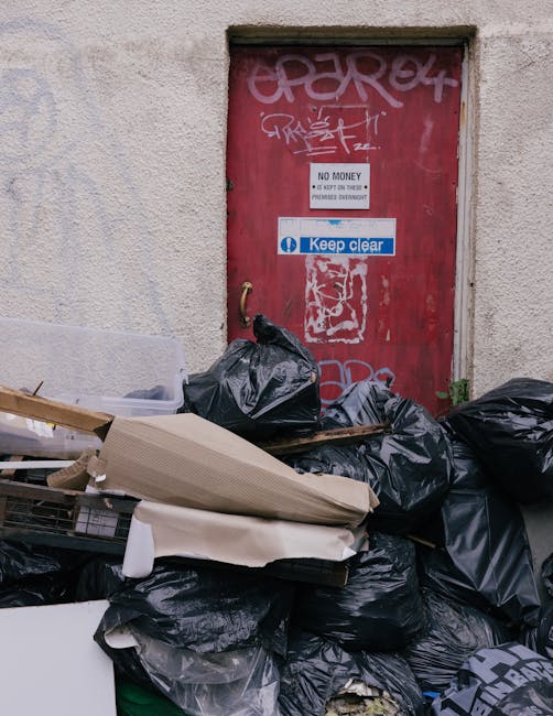A pile of black rubbish bags and discarded packaging materials stacked in front of a red, graffiti-covered door set into a concrete wall. The bags appear full and are arranged on a pavement or walkway area, with some cardboard and plastic items partially visible among the debris. The door features various stickers and graffiti tags, including a prominent blue and white sign that reads 'Keep clear,' alongside other smaller signs such as 'No Money' and instructions about access. The environment suggests an urban or alleyway setting, where independent waste collection or on-site clearance may be necessary, and the scene represents clutter typical of rubbish removal services like those provided by Rubbish Removal Maida Vale. The lighting appears natural and diffuse, without strong shadows or highlights, emphasizing the utilitarian nature of the scene related to waste handling and disposal efforts.
