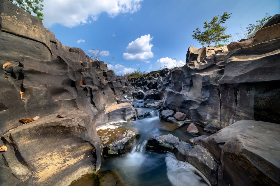 A narrow rocky gorge featuring dark, smooth-bedded stone walls with various shades of brown, gray, and black, several large, angular boulders nestled within the streambed, and a small water course flowing over and around the rocks. The water appears in motion, creating a blurred effect due to a long exposure, and the surface reflects patches of white foam. In the background, the sky is partly cloudy with bright white clouds scattered across a blue backdrop, and a few sparse trees with green foliage are visible atop the rocks on either side of the gorge. The environment suggests a natural outdoor scene with a combination of rugged rock formations and flowing water, but it lacks any visible rubbish or waste materials, aligning with natural landscape imagery rather than waste management contexts.