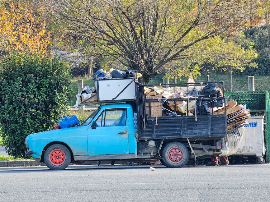 A small blue pickup truck parked on the side of a street, loaded with a large amount of mixed waste and rubbish. The cargo area is filled with cardboard boxes, plastic bags, and other debris, secured with ropes. The truck's body shows signs of wear, with chipped paint and visible rust on the red-painted wheels. It is positioned next to a neatly trimmed green hedge and a tree with sparse, yellow-green foliage, indicating an outdoor environment in an urban or suburban area. The background features a fence, some additional vegetation, and a partly sunny sky, creating natural lighting conditions that highlight the cluttered load on the vehicle. The scene reflects a type of private rubbish collection or on-site clearance often associated with alternative waste management options, with the presence of the Rubbish Removal Maida Vale company logo suggesting professional waste handling services.