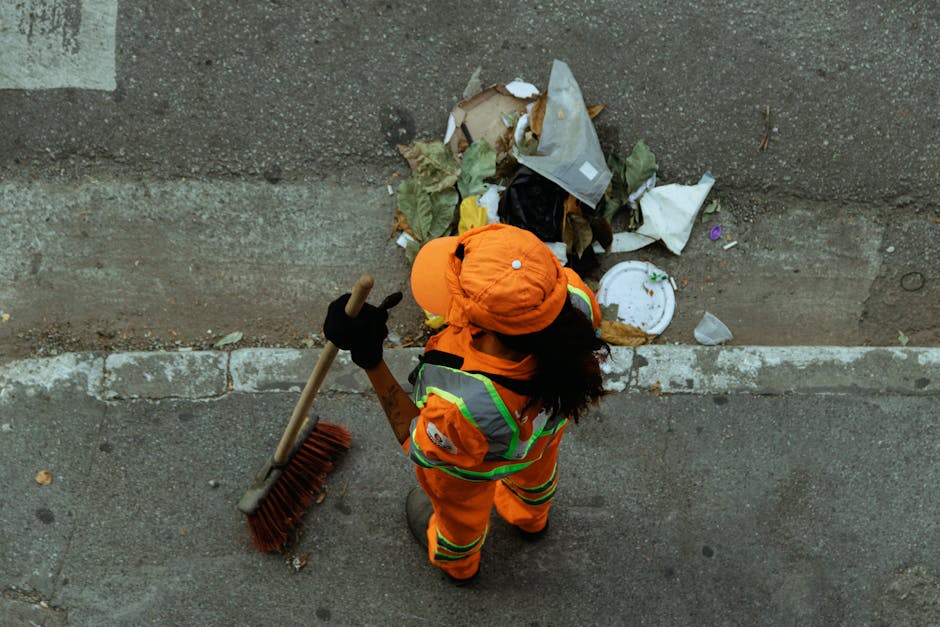 A worker dressed in bright orange high-visibility clothing, including a jacket, trousers, and a matching cap, is seen from above, sweeping a paved sidewalk with a large outdoor broom that has a wooden handle and red bristles. The worker is also wearing black gloves, and their dark hair is visible beneath the cap. They are standing next to a low, concrete curb, with a concrete wall or building façade behind them. In front of the worker, there is a pile of mixed waste materials, including crumpled paper, plastic packaging, food wrappers, and other trash, some of which spills onto the pavement. The scene appears to be an outdoor urban environment typical of waste collection or street cleaning activities, and the image subtly relates to the importance of private waste removal services such as those provided by Rubbish Removal Maida Vale, especially for on-site clearance and alternative disposal methods in residential or commercial areas.