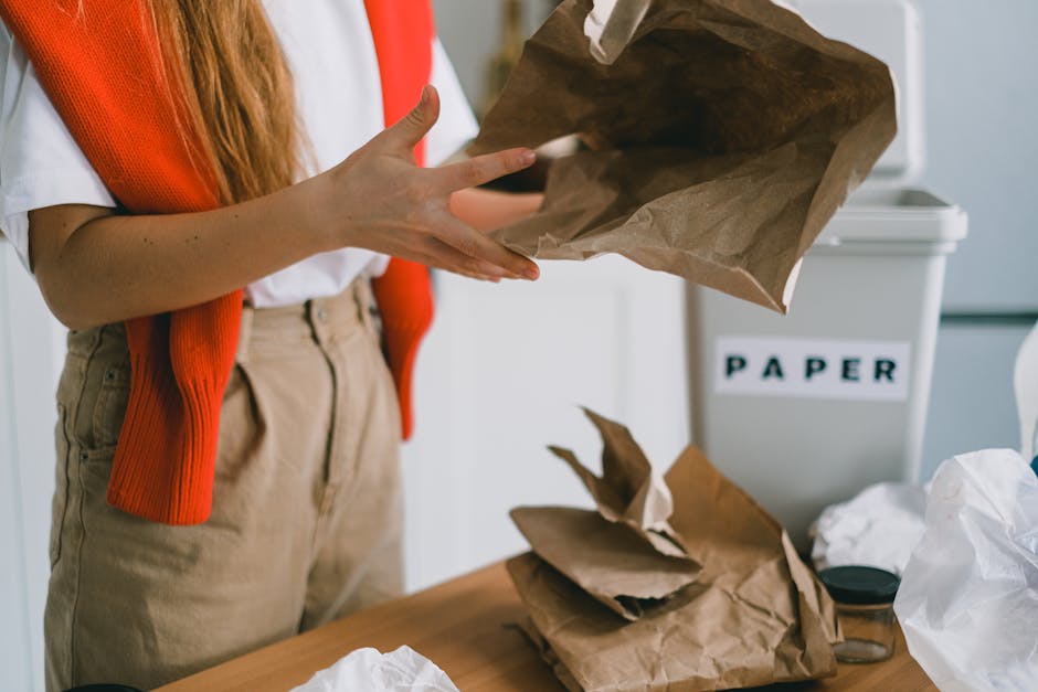 Close-up view of a person wearing a white shirt and orange cardigan, holding an empty brown paper bag with both hands, preparing it for waste disposal. The scene is set indoors, in front of a white recycling bin labeled 'PAPER' with black lettering, situated on a light-colored wooden surface. Several crumpled and torn paper bags are scattered on the table, indicating ongoing waste sorting or collection activity. The person's beige trousers and partially visible arm suggest they are engaged in an on-site waste management task, possibly part of a private rubbish removal service. The environment is well-lit, with natural or artificial light highlighting the textures of the paper bags and the details on the person's hand. This scene captures a moment of document or paper waste separation, relevant to domestic or commercial rubbish removal efforts implemented in residential settings, emphasizing the importance of proper waste handling and recycling practices in line with independent rubbish collection methods facilitated by companies like Rubbish Removal Maida Vale.