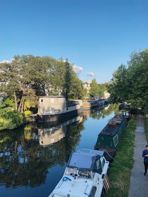 A canal scene during daytime featuring a calm waterway with several narrowboats moored along the right bank. The boats vary in size and color, predominantly dark green, black, and grey, with some displaying windows and roof fittings. On the left side, a large houseboat with a greyish exterior and rounded port-hole windows is docked close to the greenery. The canal is bordered by a well-maintained pavement and a grassy embankment, with a pedestrian walking along the path on the right. Tall, leafy trees line both sides of the canal, casting reflections onto the water's surface, which is smooth and mirror-like, capturing the shapes of the boats and trees. The sky above is clear and bright with a few scattered clouds, indicating a sunny day. This scene exemplifies an environment where private boat mooring and on-site maintenance or clearance might be relevant, and it aligns with independent waste handling or rubbish removal services in the Maida Vale area, similar to what Rubbish Removal Maida Vale offers for local residents and boat owners.