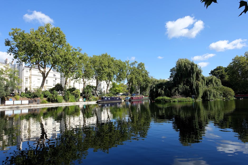 A street scene in Maida Vale features a collection of multi-storey buildings with varying architectural styles, including brick facades, arched and rectangular windows, and decorative elements. The foreground shows a white waste collection vehicle parked along the curb, associated with Rubbish Removal Maida Vale, indicating a private rubbish collection or on-site clearance service. The vehicle has a clean, modern design with a smooth, reflective surface, and is positioned adjacent to a gated entrance with stone pillars, possibly leading to a private property or courtyard. To the right, a row of motorbikes is secured along a metal railing, while further down the street, other vehicles and street furniture are visible. The background reveals additional buildings with ornate architectural details, flat rooftops, and a chimney or tower with a dome-shaped top, emphasizing the urban environment. The scene is illuminated by natural daylight, with soft shadows indicating overcast weather, highlighting the textures of bricks, glass, and metal, conveying the typical urban landscape where rubbish removal services operate to manage waste generated in residential and commercial properties.