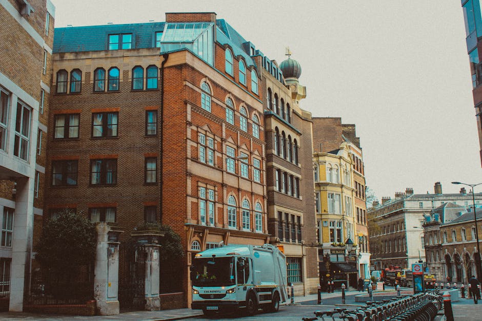 The image depicts a city street scene featuring a row of multi-story buildings with distinct architectural styles. The building in the foreground, on the left, has a brick facade with a combination of dark red and brown tones, arched window frames, and a decorative cornice beneath the roofline. Adjacent to it is a structure with a beige exterior, large rectangular windows, and an ornate bay window on the upper floor. To the right, a yellow-painted building with white trim and intricate detailing around its windows is visible. The street is lined with various elements typical of urban environments, including a white waste collection truck parked near the curb, with the logo and text of Rubbish Removal Maida Vale visible on its side. Bicycles are parked along a black metal rack in the foreground, and a few pedestrians are walking further down the pavement. Overhead, the sky is overcast, providing neutral lighting that emphasizes the textures and colors of the buildings and street furniture, illustrating a typical urban scene consistent with private waste collection and rubbish removal services in the area.