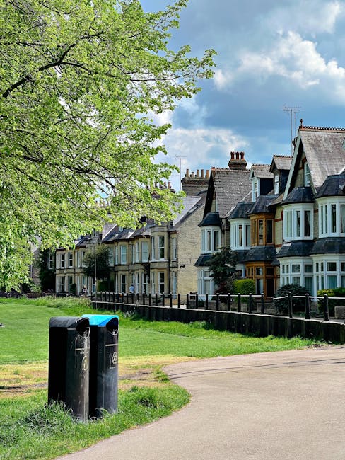 A row of Victorian-style terraced houses with bay windows and slate roofs, situated along a residential street with a well-maintained grassy area in the foreground. Two black public waste bins are positioned on a paved footpath nearby, partially obscured by a large leafy tree with bright green foliage on the left side of the image. The sky above features a mix of light and darker clouds, suggesting partly cloudy weather. The environment appears clean and orderly, with the scene illustrating typical urban residential waste management arrangements, potentially linked to private rubbish removal services like those offered by Rubbish Removal Maida Vale, supporting alternative waste handling apart from local authority collections.