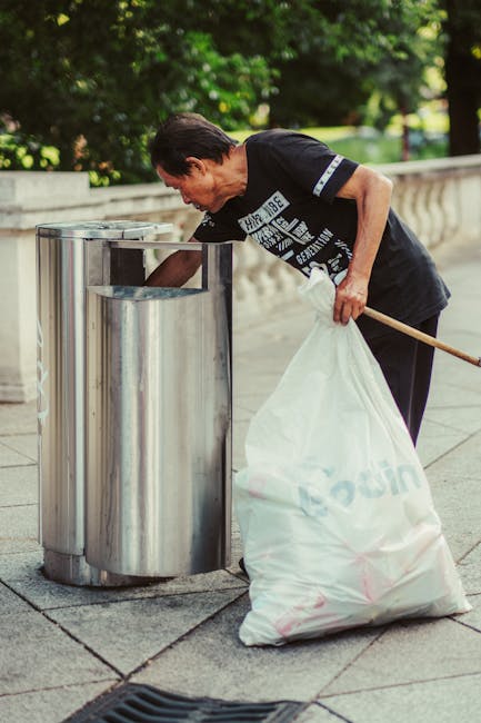 A middle-aged woman with dark hair tied back is seen disposing of rubbish into a silver, cylindrical public waste bin with multiple compartments, located on a paved pathway. She is wearing a black short-sleeved T-shirt with white printed text and dark trousers. In her right hand, she holds a white plastic bag filled with rubbish, while her left hand is lowered into the bin, which has a rectangular opening. The bin appears to be made of stainless steel, with smooth, reflective surfaces, situated near a low stone or concrete railing, and surrounded by lush green foliage and trees, indicating an outdoor park or public space. The scene is illuminated with natural daylight, casting minimal shadows, creating a clear, detailed view of her actions, which relate to waste disposal, a common task in public rubbish removal services, highlighting the importance of proper rubbish handling in community environments.