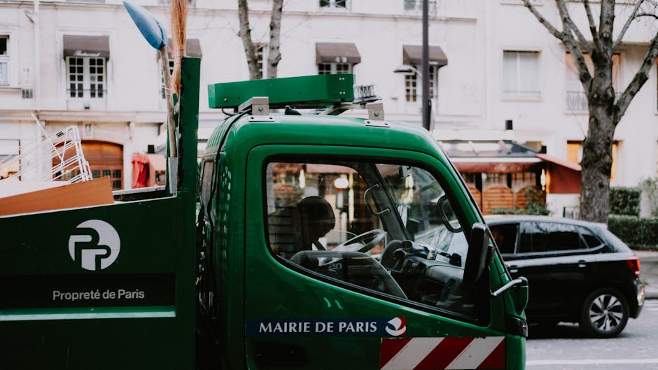 A close-up view of a green waste collection truck parked on a street lined with leafless trees and residential buildings. The truck's cab, with a clear, slightly curved windshield showing the interior steering wheel and dashboard, is in the foreground. The exterior of the truck displays the logo and text 'Propreté de Paris' and 'Mairie de Paris' on a dark blue strip, indicating its association with Paris municipal waste services. In the background, a dark-colored vehicle is parked behind the truck, and there are residential buildings with white facades, large windows, and some visible hanging laundry or coverings. The scene is captured during daylight, with natural light illuminating the scene and casting soft shadows, demonstrating the context of environmental and waste management operations typical of local authority or private rubbish collection services.
