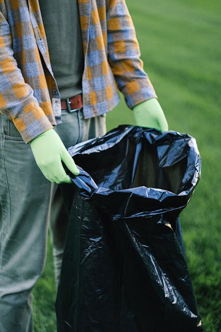 A person wearing a checked shirt with shades of yellow, blue, and gray, and green rubber gloves, is holding open a large black rubbish bag made of thick plastic. The bag appears to be partially filled with waste and is situated outdoors on a green grassy area. The individual’s lower body, including beige or light-colored trousers and a brown belt, is visible, but their face and upper body are not shown. The background features a well-maintained lawn or field with a natural, outdoor environment. The scene reflects aspects of waste collection or disposal, possibly as part of an environmental cleanup or rubbish removal activity, with the person preparing the bag for rubbish collection or disposal services offered by Rubbish Removal Maida Vale, highlighting the importance of proper waste handling and different options for waste management outside of local authorities.
