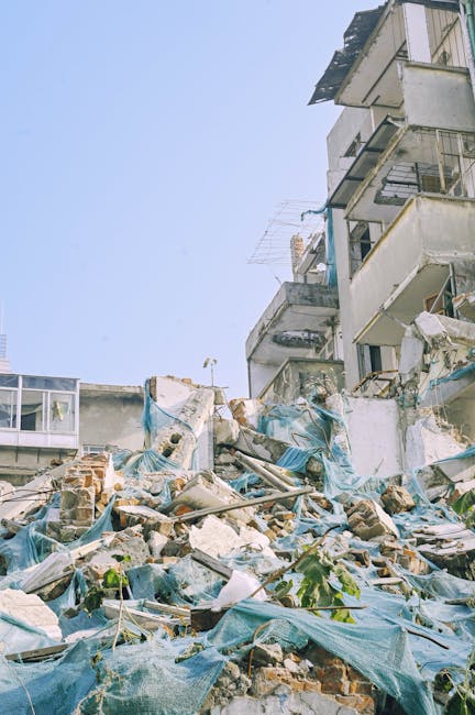The image depicts a section of a multi-storey residential building with several balconies and a weathered façade, situated against a clear blue sky. In the foreground, there is a large accumulation of construction debris and discarded building materials, including broken bricks, concrete fragments, and torn blue tarpaulins, which are partially covering the rubble. The debris appears to be loosely piled on a paved area, possibly a driveway or ground level adjacent to the building. Some green plants are visible partially emerging through the refuse, adding a contrasting natural element. The overall scene suggests an area in need of clearing or waste removal, which relates to private waste handling or on-site clearance services such as those provided by Rubbish Removal Maida Vale. The lighting is natural, highlighting the textures of the debris and the building’s exterior, emphasizing the contrast between the demolition remnants and the structure above, creating a snapshot of environmental clearing in an urban setting.