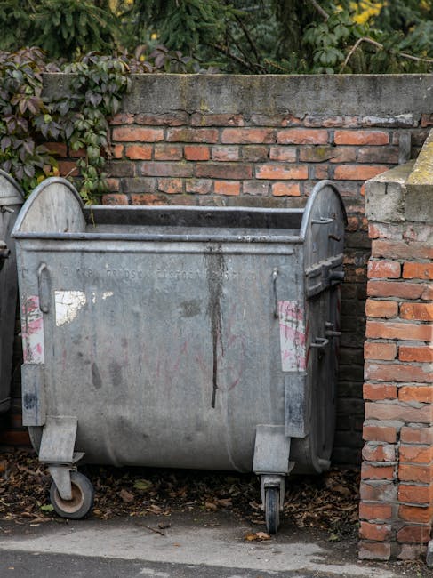 A large, metal wheeled refuse bin with a weathered, grey finish and remnants of pink and white labels on its surface, positioned on a concrete pavement against a brick wall. The bin has visible rust patches, dirt marks, and signs of wear, and features a partially open lid revealing its dark interior. The surrounding environment includes a background of green foliage and a brick wall topped with a horizontal cement coping stone, indicating an outdoor setting typically associated with waste collection points and alternative waste handling arrangements. The scene captures an everyday urban rubbish storage area often used by private waste removal services, such as Rubbish Removal Maida Vale, for on-site clearance before collection.