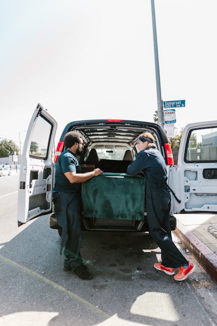 Two waste removal professionals from Rubbish Removal Maida Vale are seen loading a large, dark green upholstered sofa with a textured fabric cover into the back of a white panel van. The sofa is positioned centrally in the open rear door of the vehicle, with one worker on each side holding the piece, suggesting coordinated lifting or manoeuvring. The van is parked on a paved street, with the open pavement and curb visible in the foreground. In the background, a street sign, a light pole, and some trees can be seen, indicating an urban environment. Bright daylight illuminates the scene, creating clear shadows on the ground and highlighting the texture of the sofa’s fabric. The scene reflects a private waste clearance activity, typical of independent rubbish collection services aimed at removing bulky items that require special handling outside standard council collection, with the focus on responsible and professional disposal of large household refuse items.