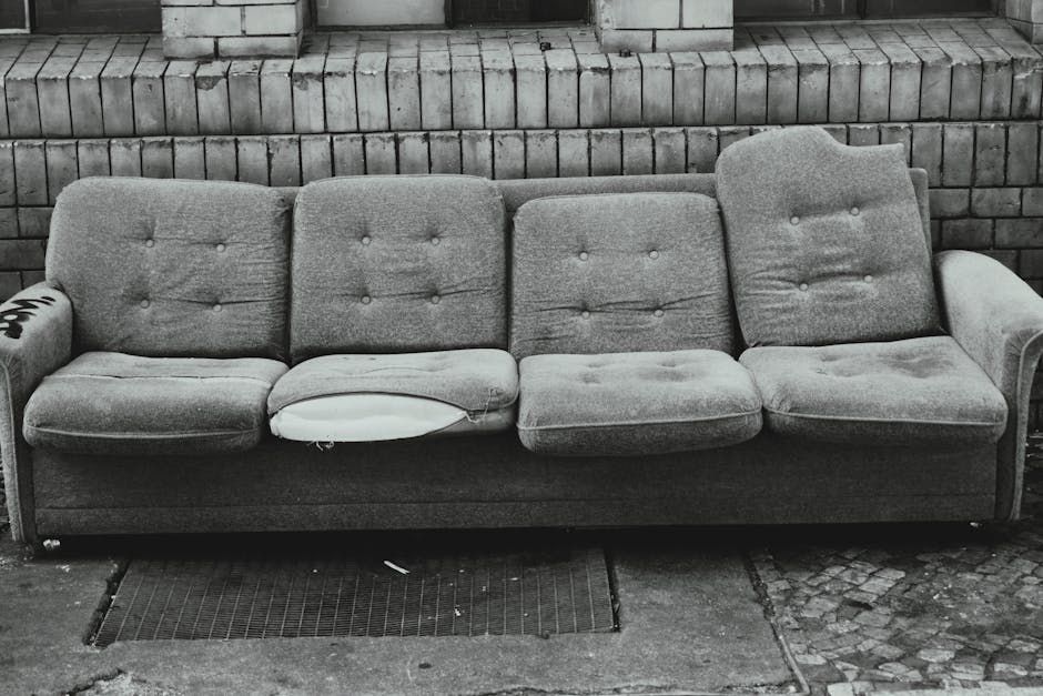 A weathered grey fabric sofa, positioned outdoors against a brick wall, with visible signs of wear including torn and sagging upholstery, especially on the middle seat where the fabric is torn, exposing the cushioning. The sofa features button-tufted back cushions and rounded armrests, with one of the armrests partially ripped and showing foam padding inside. It is situated on a paved surface that includes a metal drainage grate directly beneath the front edge of the sofa, indicating an exterior environment such as a driveway or pavement. The background consists of a horizontally laid brick wall with variations in tone, suggesting an aged or unmaintained setting. The sofa's appearance and the surrounding environment imply that it has been discarded and is awaiting collection, possibly through private waste removal services like Rubbish Removal Maida Vale, to facilitate on-site clearance or alternative waste disposal.