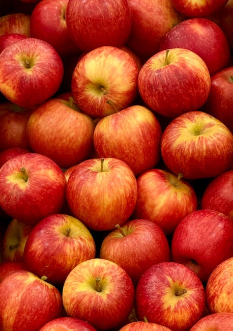 A close-up view of multiple fresh apples piled together, featuring round, smooth surfaces with a blend of red and yellow hues. The apples display natural variations in coloration, with some showing streaks and patches of lighter yellow-green near their stems. The skin of the apples has a slightly shiny finish, reflecting light that enhances their vibrant appearance. The apples are arranged tightly, occupying the entire frame, with some overlapping each other. The background is not visible, focusing solely on the uniform cluster of apples, which appear to be in a natural setting or display. This image emphasizes the freshness and vivid coloring of the fruit, commonly associated with produce stores or grocery sections, subtly supporting themes of food quality and freshness related to waste reduction in food use.